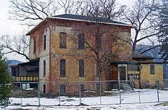 A two-story brick house in extremely poor condition, with boarded-up windows and extensively discolored white paint. It is fenced off.