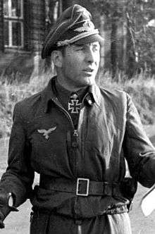 Black-and-white portrait of a man wearing a peaked cap and military flight suit with an Iron Cross displayed at his neck.