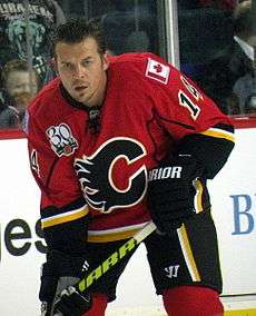 Hockey player in red uniform with a big "C" in the middle. He stares, grim faced, directly at the camera, his stick pointing downward