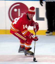 A hockey player in a red uniform with white and yellow trim.  He is focused on the ice surface, as he attempts to control a puck while skating