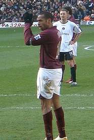 Thierry Henry, wearing gloves and a redcurrant football shirt applauds the crowd. A stand full with people and man wearing a football shirt are visible in the background.