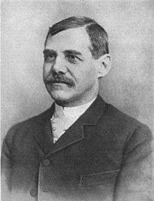 A man with dark hair and a mustache wearing a high-collared white shirt and a black jacket