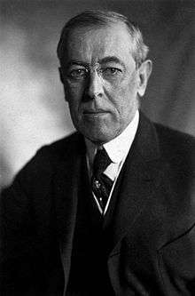 Half-length portrait of a man wearing a three piece suit, a neck tie, round rimmed glasses and looking directly towards the photographer with an emotionless expression.