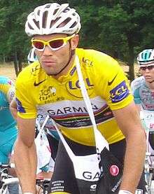 A road racing cyclist wearing a yellow jersey with a white section on the chest, which contains rainbow stripes as well. He has a musette bag on over his head, and other cyclists are visible in the background.