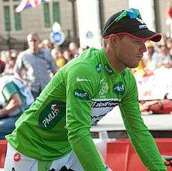 A man, presumably sitting on a bicycle, wearing a green jersey and a black cap with sunglasses on. In the background one can spot spectators.