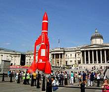 A model space rocket stands ready for lift-off in a crowded city square