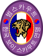 An oval badge of red with 범스카우트 (Tiger Scout) and 한국 보이 스카우트 연맹 inscribed in hangul; The center is an oval of yellow with the profile of a tiger head over a scroll inscribed with "preparation" and knot