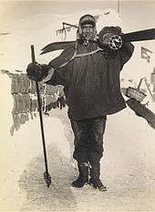  Man, standing, wearing a smock, heavy trousers and boots. He has a ski stick in his right hand, a pair of skis strapped on his back, and is carrying a rounded bundle on his shoulder. Behind him on the ground is assorted polar equipment.
