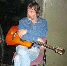 Color photograph of a casually seated middle-aged bearded male wearing casual clothing and resting a guitar on his lap.