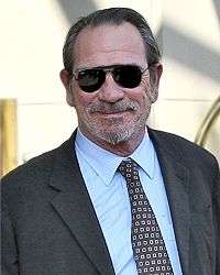 A man with dark hair and a short beard wearing sunglasses and a grey suit with a patterned tie.