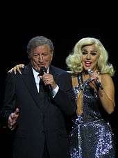 Bennett in suit on the left and Gaga in a sparkling dress and blonde hair singing onstage
