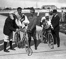 Two men with flowers in their hands, sitting on bicycles being held by other men, with the Eiffel tower seen in the background.
