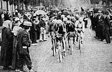 A group of cyclists, with spectators on the left and on the right.
