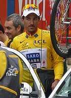 A cyclist in a yellow jersey enters a car with a bicycle on top of it. Other people are visible behind him.
