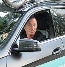 A man in his fifties behind the wheel of a car, as seen from the driver's side. He is looking out the opening left by the rolled-down window, to the left of the car.