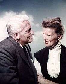 Hepburn is sitting with Spencer Tracy, she age 50 and he age 57, and they are smiling at each other.