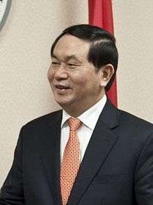 a smiling man with black hair, dressed in a suit with an orange tie