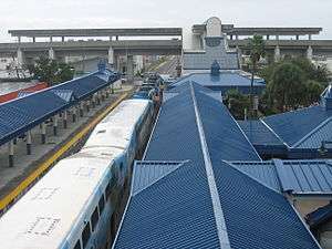 A commuter train station with blue roofs featuring a large train on one of the tracks.  A connecting elevated metro station is in the background.