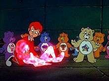 A boy dressed in red looks down at a girl with a crystal glow. A yellow bear stands up with her hands outstretched; some of her partners are holding hands and chanting.