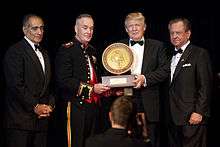 A ceremony in which Trump receiving the 2015 Marine Corps–Law Enforcement Foundation's annual Commandent's Leadership Award. Four men are standing, all wearing black suits; Trump is second from the right. The two center men (Trump and another man) are holding the award.