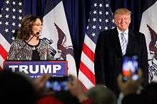 Donald Trump with former Alaska governor Sarah Palin in January 2016. Palin is standing on the left side of the image, behind a podium with a sign that has the word "TRUMP" in white-on-blue text. Trump is standing on the right side of the image. There are American flags hanging on poles behind them and the outlines of an audience in front of them.