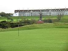 A wide, sprawling golf course. In the background is the Turnberry Hotel, a two-story hotel with white facade and a red roof. This picture was taken in Ayrshire, Scotland.