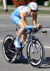 A road racing cyclist in a blue, gold, and white skinsuit, sitting low in a crouch on his bicycle.