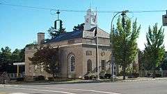 A grayish-brown stone building with a peaked roof topped by a white cupola and small trees on the side, seen from its left across an intersection with traffic lights dangling from a cord stretched across the top of the image.