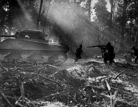 U.S. soldiers advancing at dawn through a jungle in Bougainville, behind a M4 Sherman