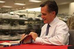 Casual photograph of Mitt Romney indoors seated and signing books