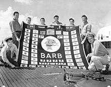 Black and white photo of eight men in military uniforms holding a large banner with the word "BARB" in the center surrounded by Japanese and Nazi flags, symbols designating military medals and symbols signifying bombardments