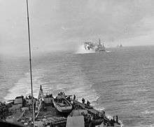 Black and white photo with part of a ship in the foreground and flames and smoke risking from the left of a warship in the background. Several other ships are visible on the horizon at the rear of the photo.