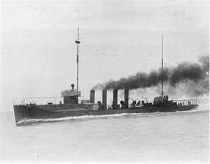 A black-and-white photo of the warship. Shown in motion with black smoke billowing from its four smokestacks, the ship has two masts but no sails.