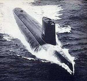 A submarine is running on the surface of the water at high speed, as evidenced by the long white wake around and behind the hull