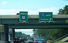 A four lane divided highway passing under a freeway. A set of Township green signs are attached to the freeway overpass, with the sign on the left reading north U.S. Route 9 and the sign on the right reading Interstate 195 west Trenton with an arrow pointing to the upper right.