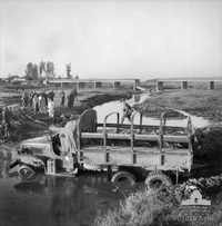 A truck sits imobile in mud and water, while soldiers and civilians look on. In the background is a damaged bridge.