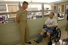 Color photograph of a standing man in uniform (left) and man in wheelchair with casts on right foreleg and left forearm (right).