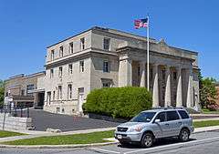 A light tan stone building with a classical colonnade and pediment on the front with an American flag flying against a blue sky from a flagpole out front. Parked on the street in front is a light blue Honda minivan.
