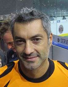 Man with brown eyes and short gray hair, wearing an orange shirt with black-lined collar.