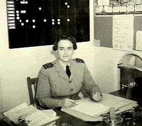 Dark-haired woman in light-coloured military uniform sitting behind a desk