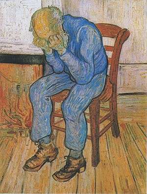 An old man with a bald head is sitting on a yellow chair by his fire. There is a low fire in the grate. He is dressed in blue clothes. He is holding his head in his hands.