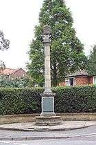 tall, thin gothic revival memorial.  A tall round pillar on a square base in a residential area, a bungalow discretely behind a hedge.  Behind the pillar is a neatly clipped hedge and a tall, green conifer.