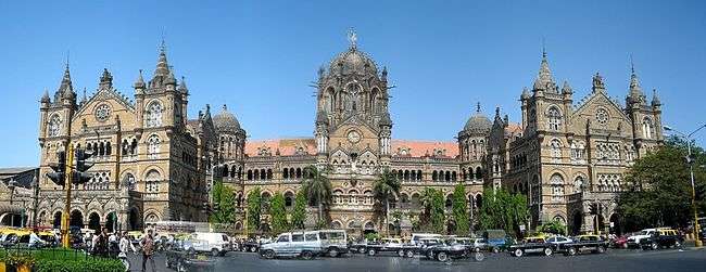 A brown building with clock towers, domes and pyramidal tops. Also a busiest railway station in India. A wide street in front of it