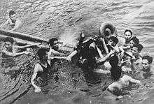 Ten men in water, surrounding one man being floated on wooden poles and tires