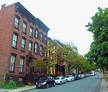 A row of three-story rowhouses in varied colors along a street with small trees and cars parked in front of them, looking uphill