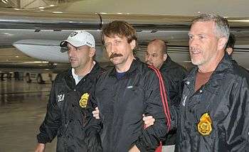 A mustached man, looking at the camera, in a shiny black warmup jacket with red stripes walking between two men wearing black jackets with large gold badge-shaped patches, and white lettering on their sleeves and breasts. It says "DE" on the man on the right and "DEA" on the man on the left. Both men are holding the adjacent elbow of the man in the center. The man on the left is wearing a white baseball cap; part of an airplane and the walls of a hangar are visible in the background