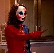 A figure wearing an outfit that is half red and half blue. The figure is wearing a mask that is split between a more masculine half and a more feminine half. The figure is pointing a knife towards an unknown target.