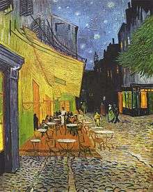 The outdoor terrace of a cafe with several tables filled with patrons. People are walking along the street under a starry sky.