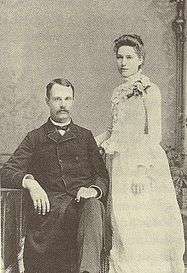 A black-and-white photograph of a couple. On the left is a seated man with a moustache weraing a dark suit. Standing on the right is a woman in a white dress, body facing left.  Both face the camera.