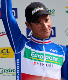 A man in his mid-thirties, wearing a cycling jersey with blue, white, and red horizontal stripes, and a black baseball cap. He is holding his right arm in the air.